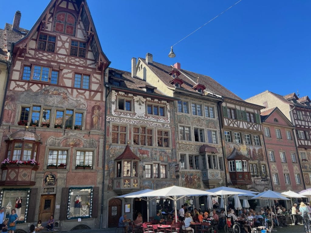 Colorful historic buildings with intricate frescoes in a European cityscape, bustling outdoor cafe scene under clear blue sky, capturing charming architecture and lively street atmosphere in Germany.