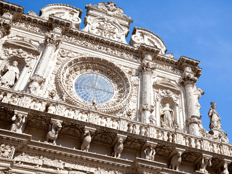 Ornate Baroque-style facade of a historic European church with carved statues and intricate detailing against a clear blue sky, showcasing architectural grandeur and religious artistry.