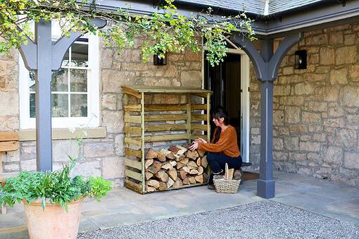 A woman stacking firewood under a covered stone porch of a rustic home with garden decor.