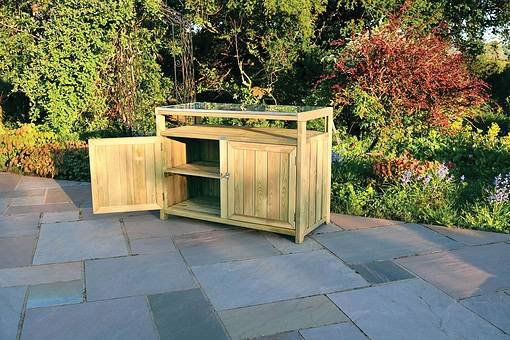 Natural wood outdoor garden storage cabinet with glass tabletop on a stone patio amidst lush garden greenery.