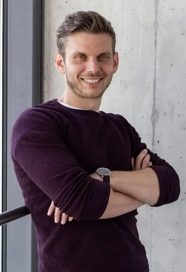 Tobias Roppelt stands with his arms crossed in front of a concrete wall and smiles at the camera. He has short, straw-coloured hair, is wearing a wine-red jumper and has rolled up his sleeves to mid-forearm.