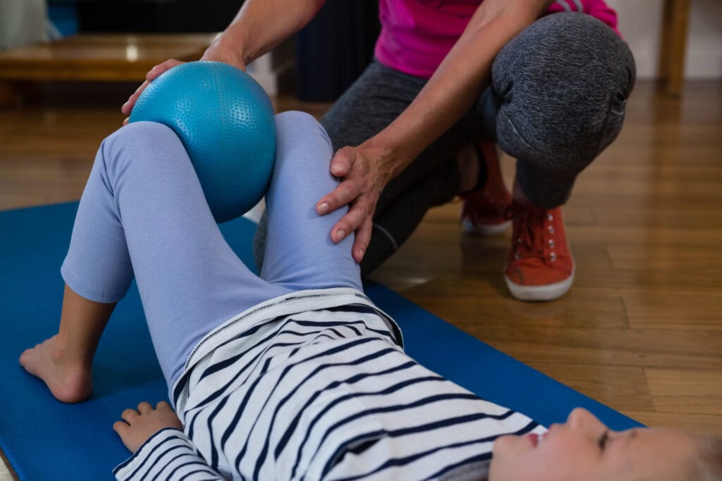 Female physiotherapist helping girl patient in performing stretching  exercise on exercise mat