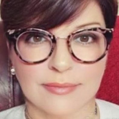 A person with short dark hair wearing large round glasses sits and looks directly at the camera.