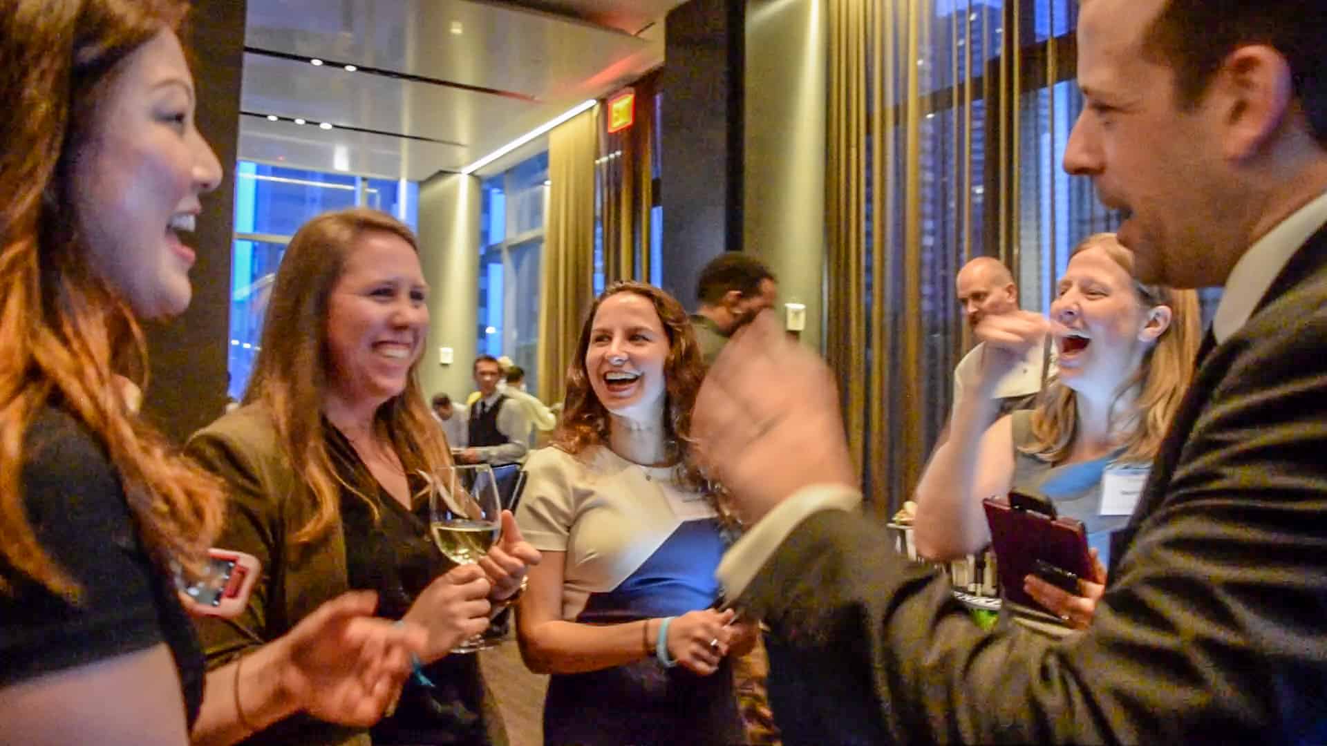 A group of five people are standing and laughing together at a social gathering. A man in a suit gestures animatedly, while two women hold drinks. The background shows a modern interior with large windows.