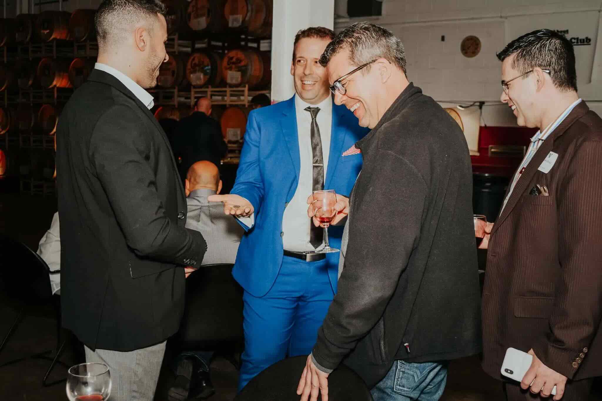 Four men are standing and talking at a social event. Three are holding drinks, and one is holding a phone. Barrels and dim lighting are visible in the background.