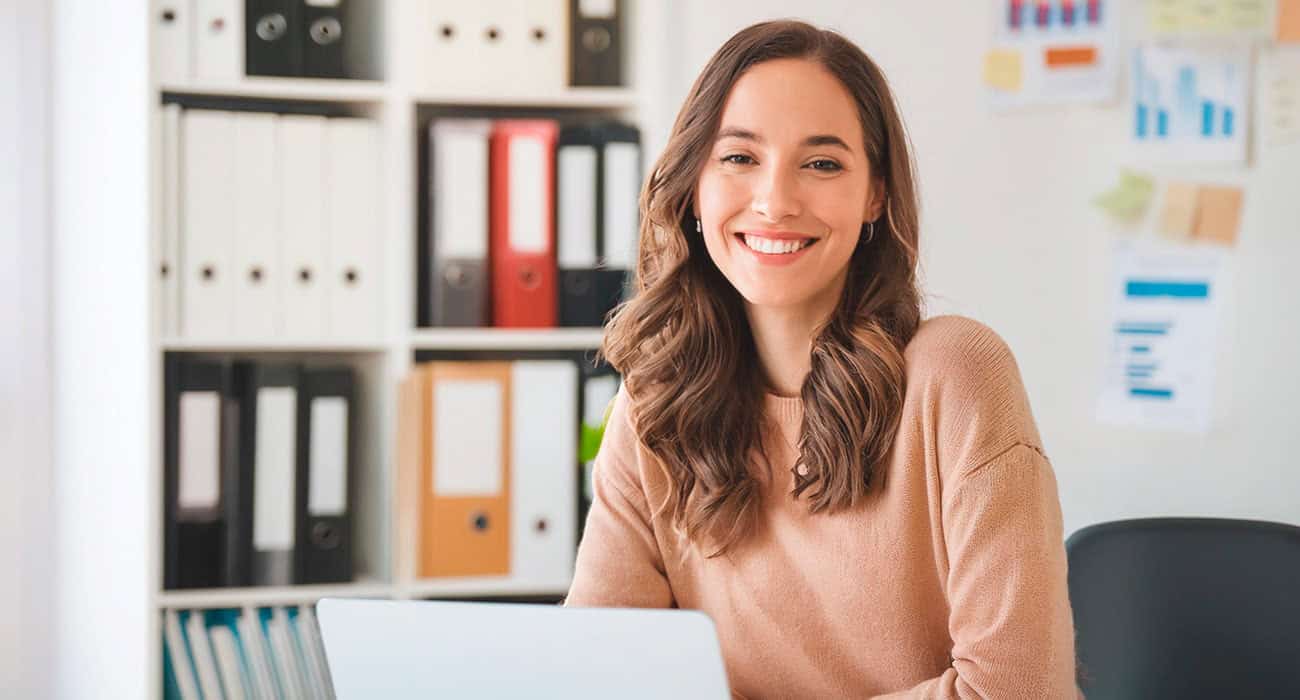 freundliche junge Dame lächelnd leger angezogenam am schreibtisch vor laptop im büro, schrank mit aktenordner unscharf im Hintergrung, helle Büromöbel
