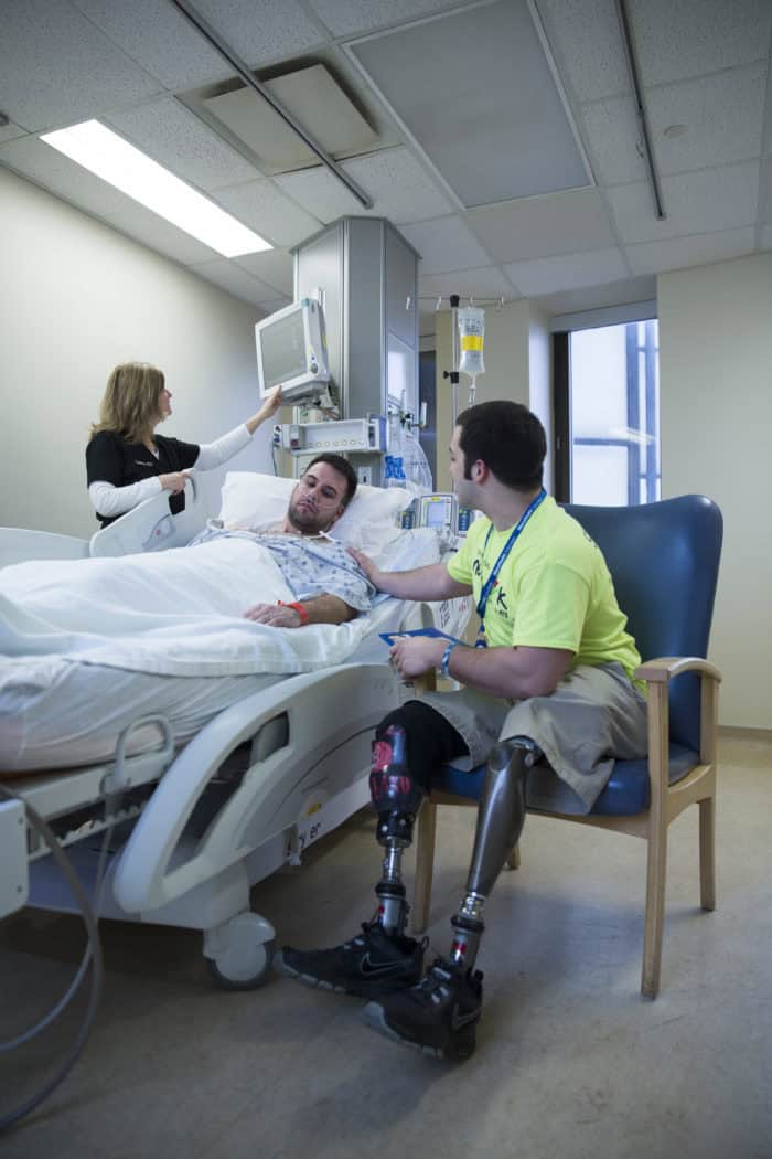 A man with prosthetic legs sits by a man in a hospital bed.