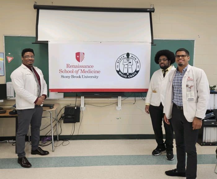 Three med school students in white coats stand by a sign with the Renaissance med school and Black Men in White Coats logos