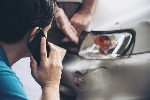 Person on phone discussing car repair near damaged vehicle.
