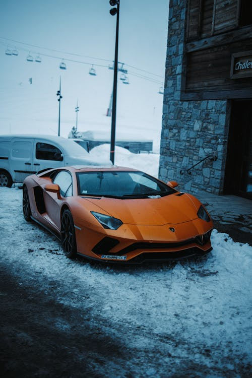 Orange Lamborghini Aventador parked on snowy ground outside a mountain lodge.