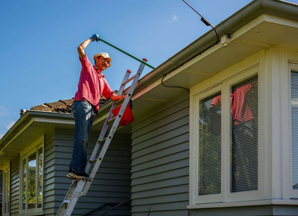 Man standing on the ladder and cleaning gutter with a gutter cleaning shovel. Home maintenance work. Auckland. | Ever Clean Clog-Free Gutter Systems - Kentucky, Cincinnati, & North Carolina