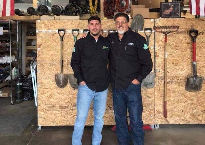 Two people in black shirts stand in a workshop with shovels and tools mounted on a plywood wall behind them.
