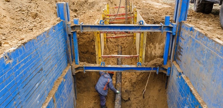 A worker installs pipes in a trench supported by steel frames.