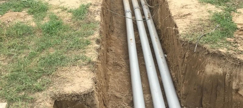 Three metal pipes installed in a dug trench beside grassy soil.