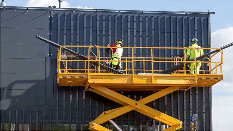 Two construction workers are performing maintenance work on the building's facade on a scissor lift with a double extension platform