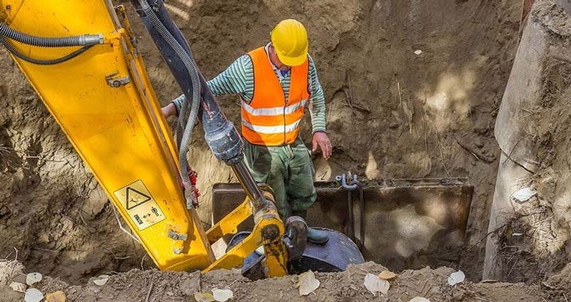 Worker in safety vest and yellow helmet working in a trench beside a backhoe.