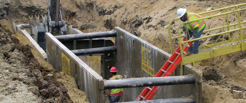 Construction workers in safety vests and helmets working in a shored trench with a red ladder.