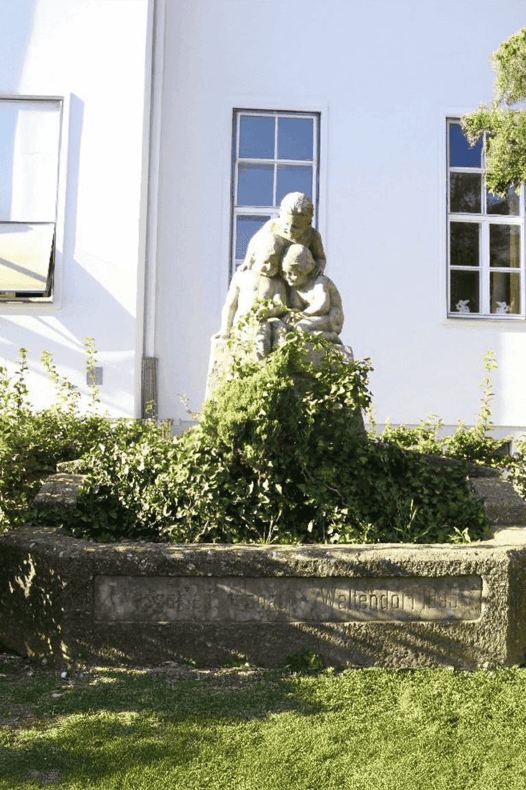 Froschkönigbrunnen in Graz-Waltendorf - stone sculpture with three childlike figures on a fountain basin, surrounded by ivy and green plants, in front of the primary school Waltendorf.