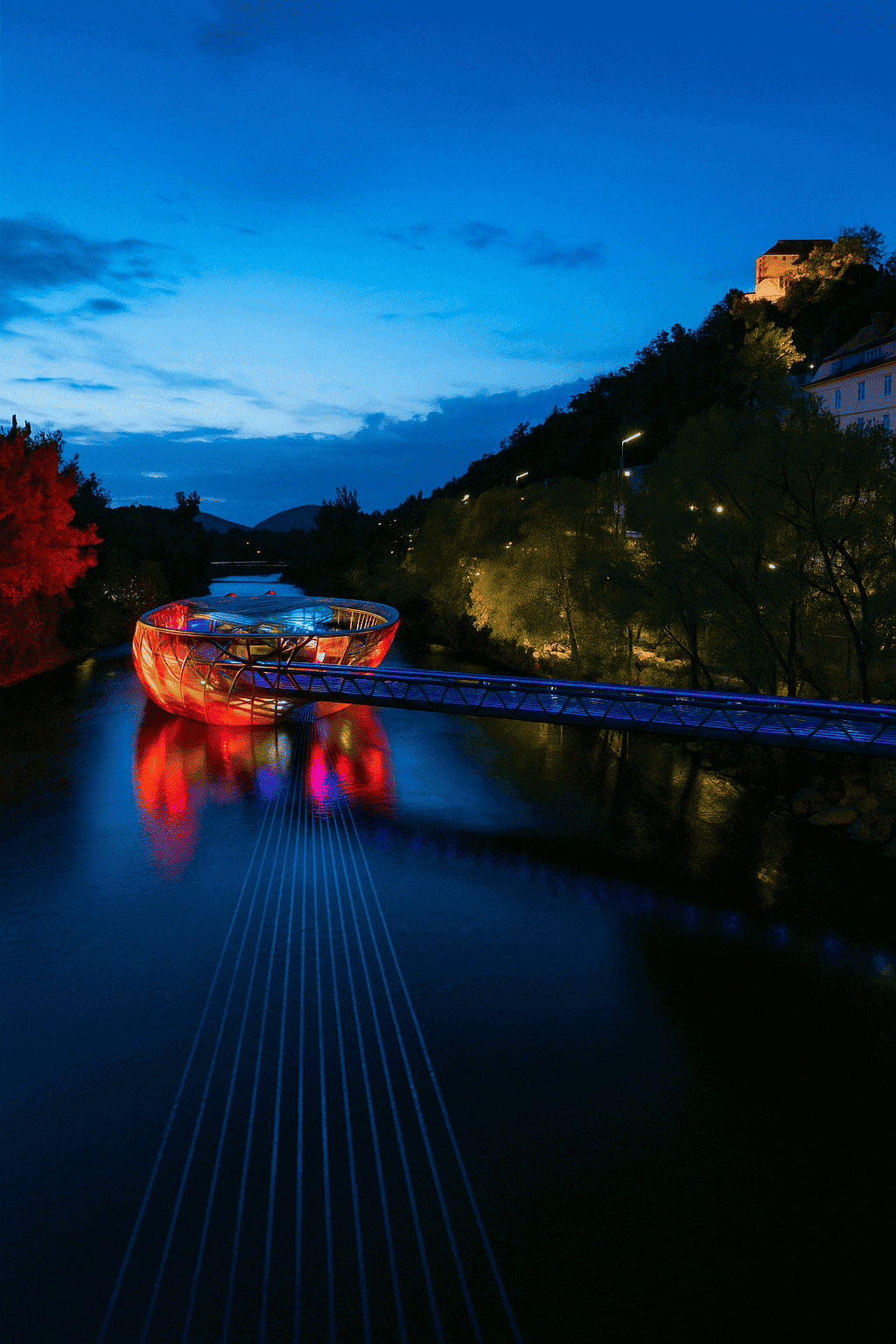 Illuminated Mur Island in Graz at night, with coloured play of light on the glass construction, reflecting in the dark water of the Mur, surrounded by trees, city lights and the Schlossberg in the background