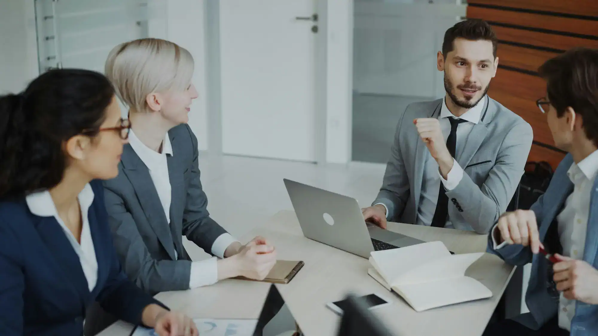 A group of four business professionals in an engaged discussion around a meeting table with a laptop and notebooks, representing the strategic partnership conversations and results-focused collaboration that define how Feedback ASAP works with client organisations.