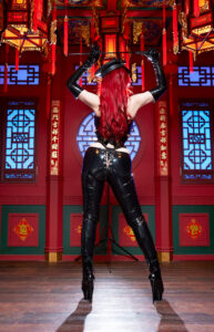 中華風の豪華な内装の部屋で、艶やかなチャイナドレスをまとい、鞭を手に堂々とポーズを決める女王様。BDSMの支配者としての威厳と官能を湛えた姿。