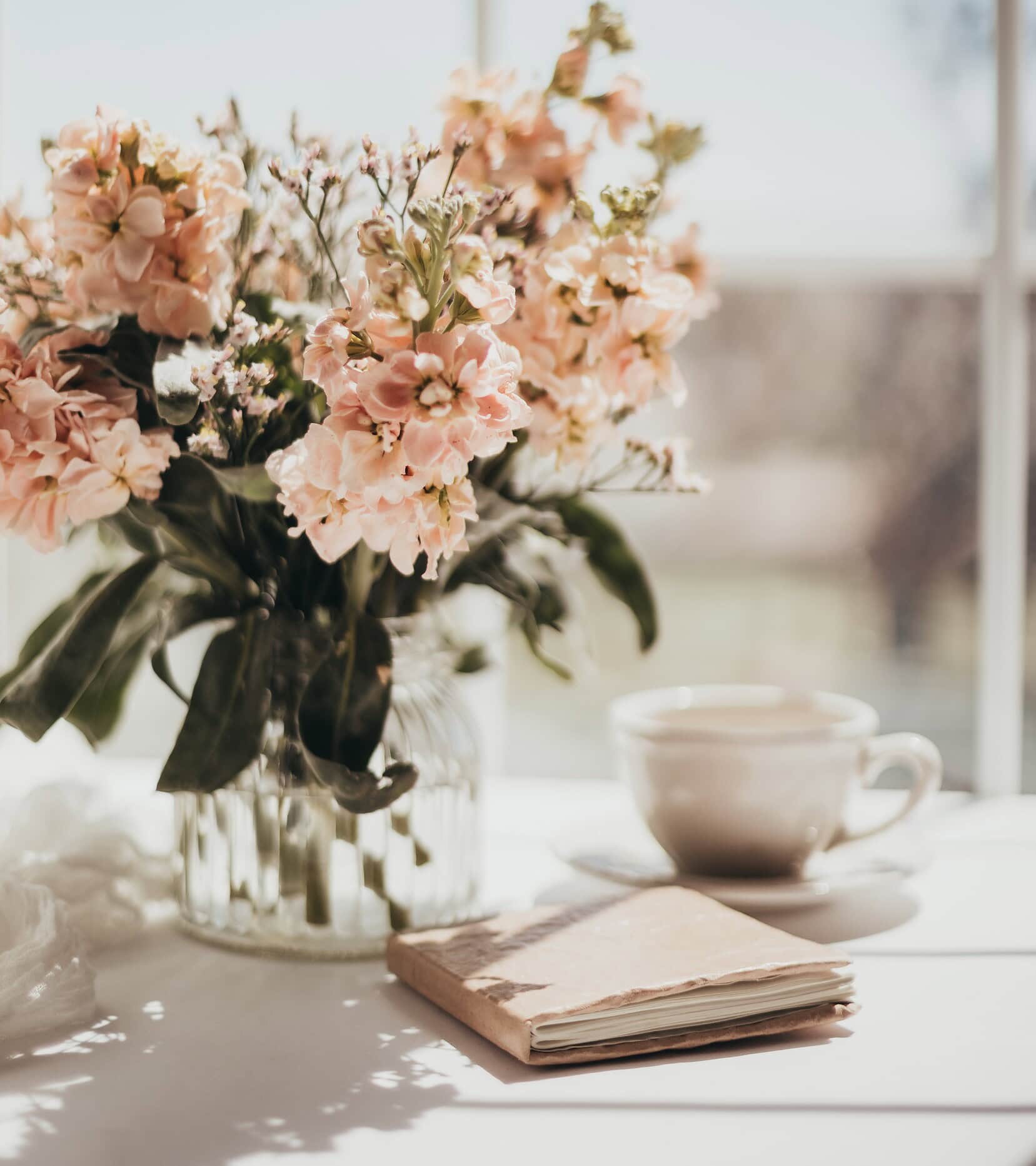 Florero con flores rosadas y taza de té en mesa soleada.