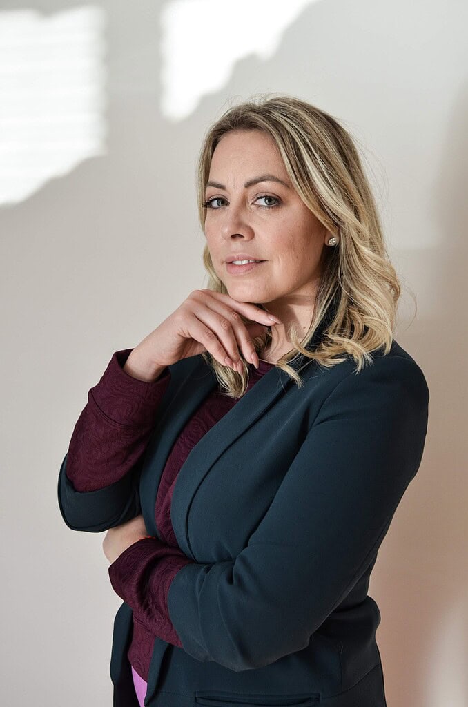 Rostro de mujer profesional con expresión confiada, cabello rubio y ropa elegante en ambiente lumino.