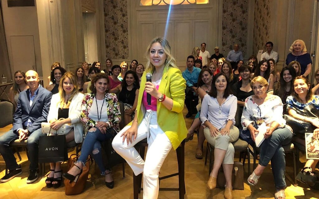 Mujer con micrófono en evento con audiencia en sala elegante y decorada.