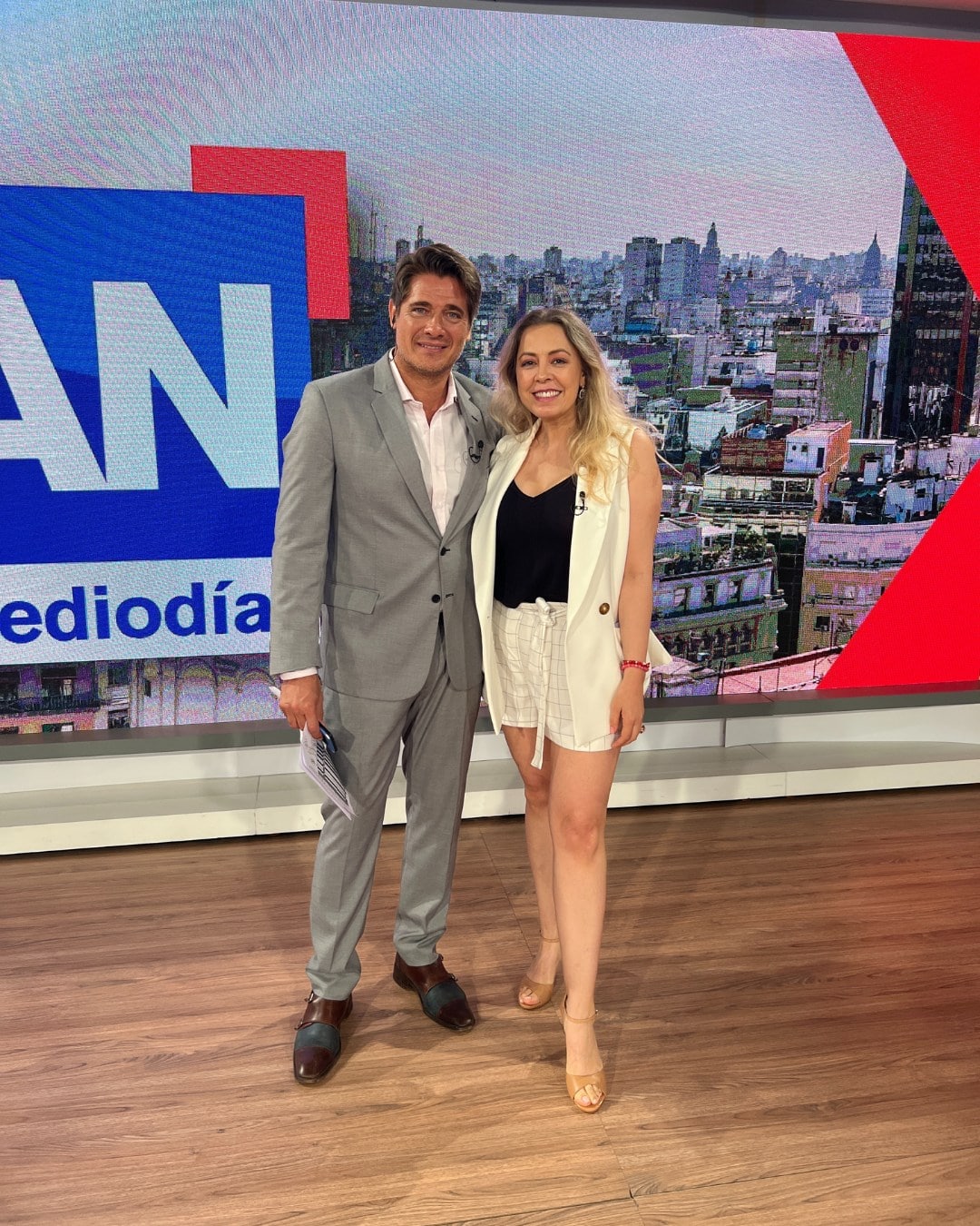 Presentadores en estudio de televisión con fondo urbano y logo en pantalla.