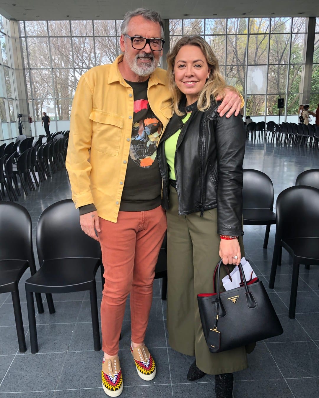 Pareja sonriente en evento con fondo de ventanas y sillas negras.