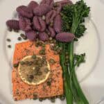 Picture of grilled salmon on a white plate paired with purple sweet potato gnocchi and baby broccoli, topped with capers.