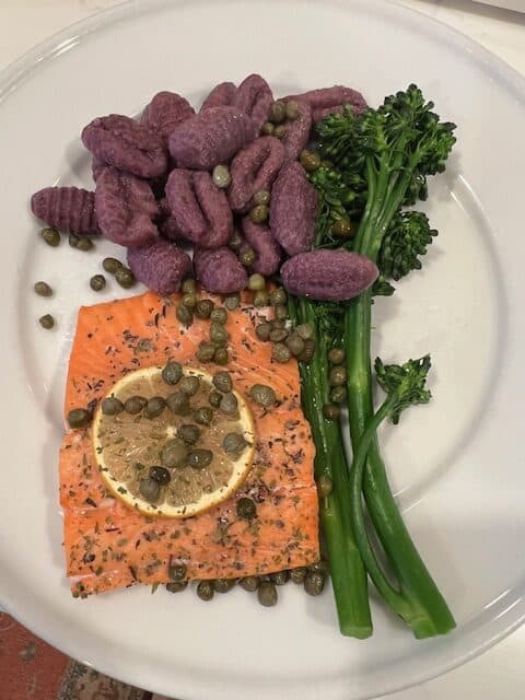 Picture of grilled salmon on a white plate paired with purple sweet potato gnocchi and baby broccoli, topped with capers.