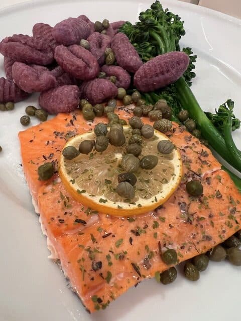Picture of a grilled sockeye salmon filet paired with purple gnocchi and baby broccoli, topped with capers. 
