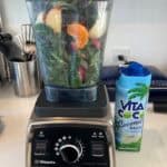 Picture of pre-blended kale & Citrus green juice loaded in a Vitamix ready to blend with a bottle of coconut water next to the blender.