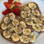Picture of two slices of sourdough bread, topped with almond butter and banana slices drizzled with honey and chia seeds, plated with strawberries to the side.