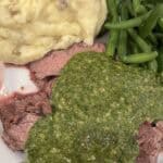 Picture of flank steak on a plate with mashed potatoes, green beans, and chimichurri sauce.