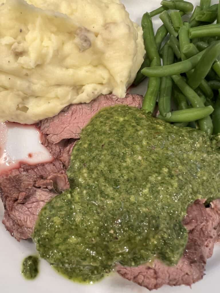 Picture of flank steak on a plate with mashed potatoes, green beans, and chimichurri sauce.