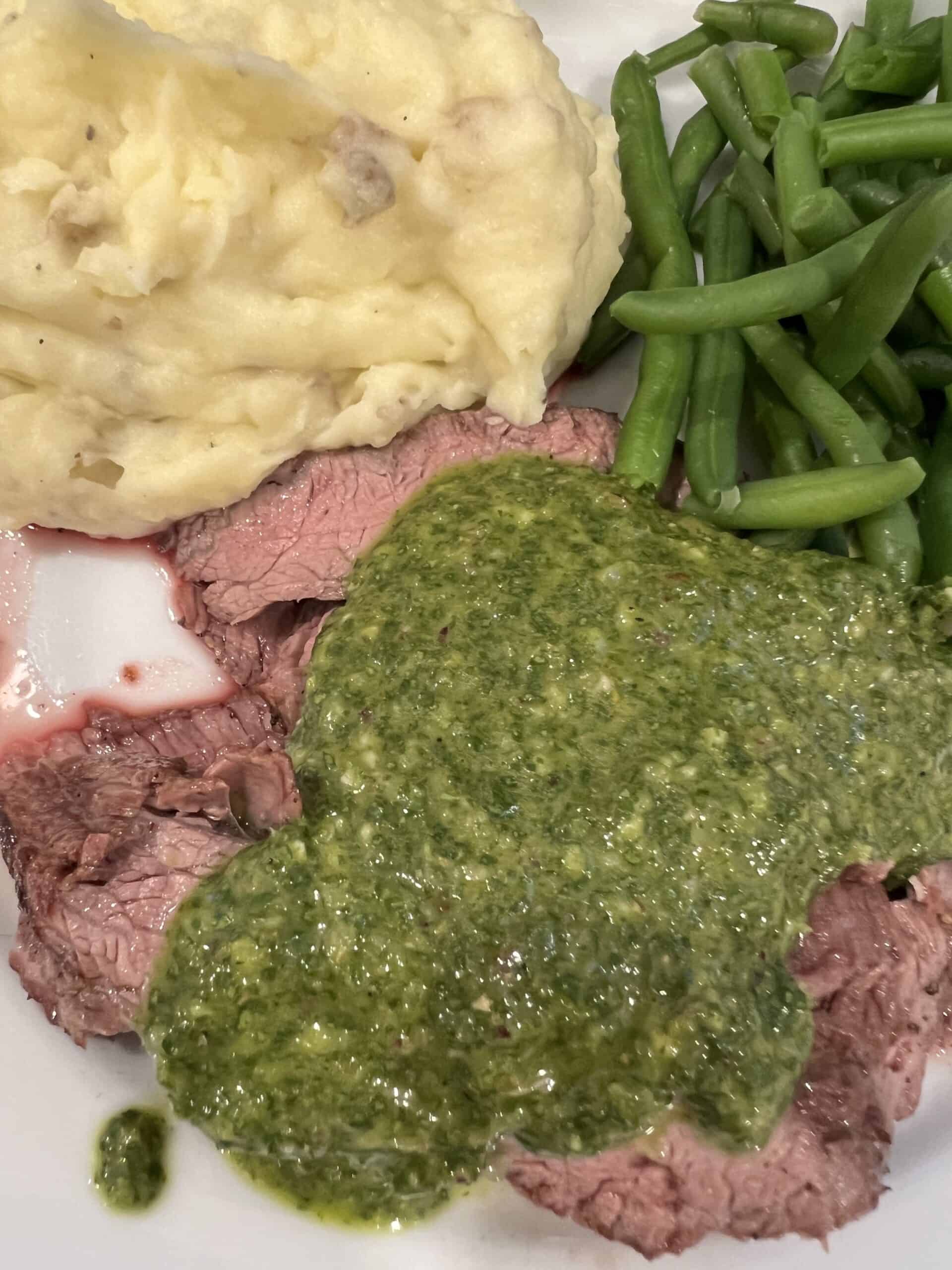 Picture of flank steak on a plate with mashed potatoes, green beans, and chimichurri sauce.
