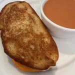 Picture of an Heirloom Tomato Gooey Grilled Cheese sandwich on a plate paired with a bowl of tomato soup.