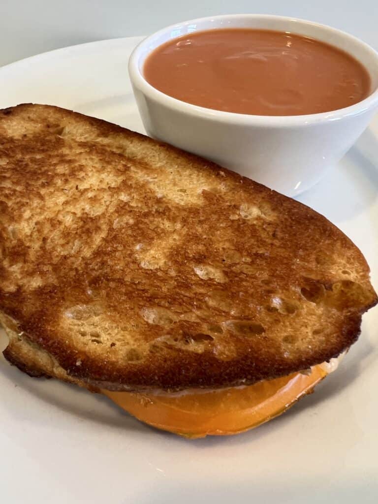 Picture of a Heirloom Tomato Gooey Grilled Cheese Sandwich on a plate paired with a bowl of tomato soup.