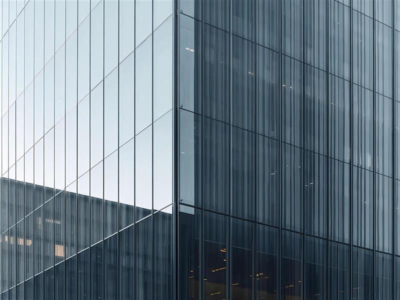 A modern glass office building with reflective windows, showing a corner where two facades meet. The building reflects the sky and nearby structures, creating a sleek and minimalist appearance.