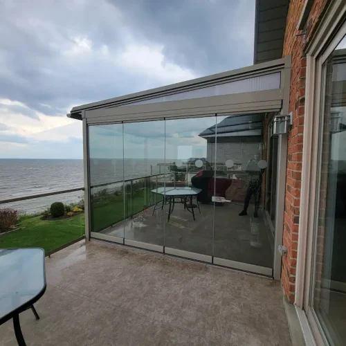 A glass-enclosed patio with a view of the sea, a table inside the enclosure, and a cloudy sky overhead. The patio is attached to a brick house with a glass door.