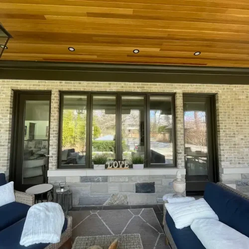 A cozy patio with cushioned seating, throw blankets, a small table, and a rug. Behind the furniture is a window with a decorative LOVE sign and potted plants. The ceiling features warm wooden paneling.