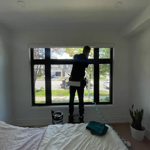 A person stands on a step stool by a large window in a bedroom, cleaning the glass. Daylight streams in, showing a street view outside. A bed, a plant, and some cleaning supplies are visible in the room.