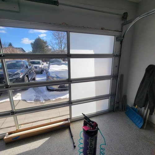 A garage with a partially frosted glass door; snow-covered cars are visible outside. Inside, there’s a blue snow shovel, some tools, a coat hanging, and a purple air compressor with a coiled hose on the speckled floor.