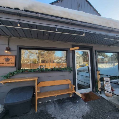 A small gray building with large windows, holiday garland on the railing, a wooden bench, and snow on the roof and ground. A sign on the left reads MARK IV BROKERAGE. A OPEN sign is by the entrance.