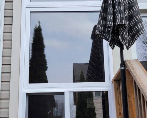 A large white-framed window reflects trees and houses. Next to it, a black patterned patio umbrella is partially open, standing beside a wooden railing and staircase. The siding of the house is beige.