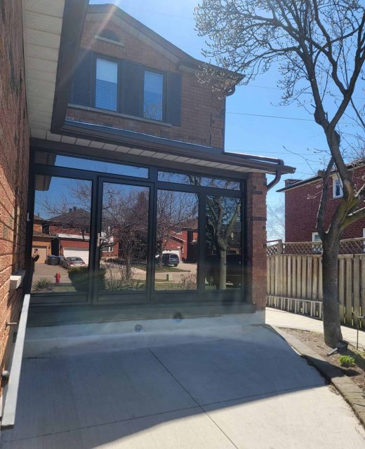 A sunlit house with large glass windows and a sloped concrete driveway. A leafless tree stands nearby, and other houses are visible in the background under a clear blue sky.