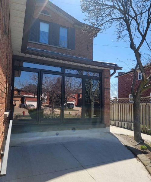A glass-walled sunroom is attached to a brick house, with a paved driveway leading up to it. Leafless trees and neighboring houses are visible under a clear blue sky.