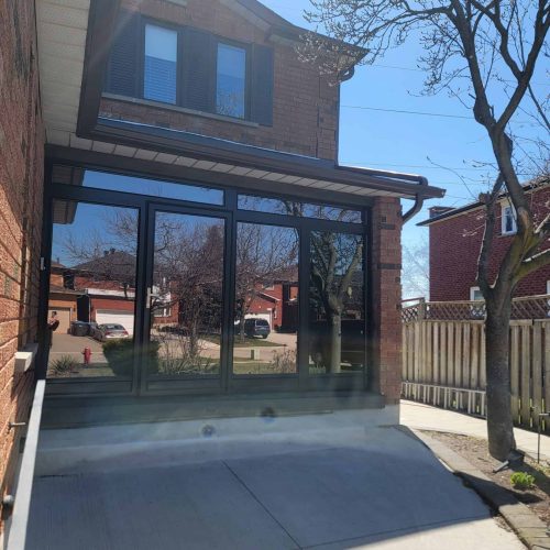 A newly built glass sunroom with black frames is attached to a brick house. The driveway is concrete, and a tree with bare branches stands nearby. Another brick house and clear blue sky are visible in the background.